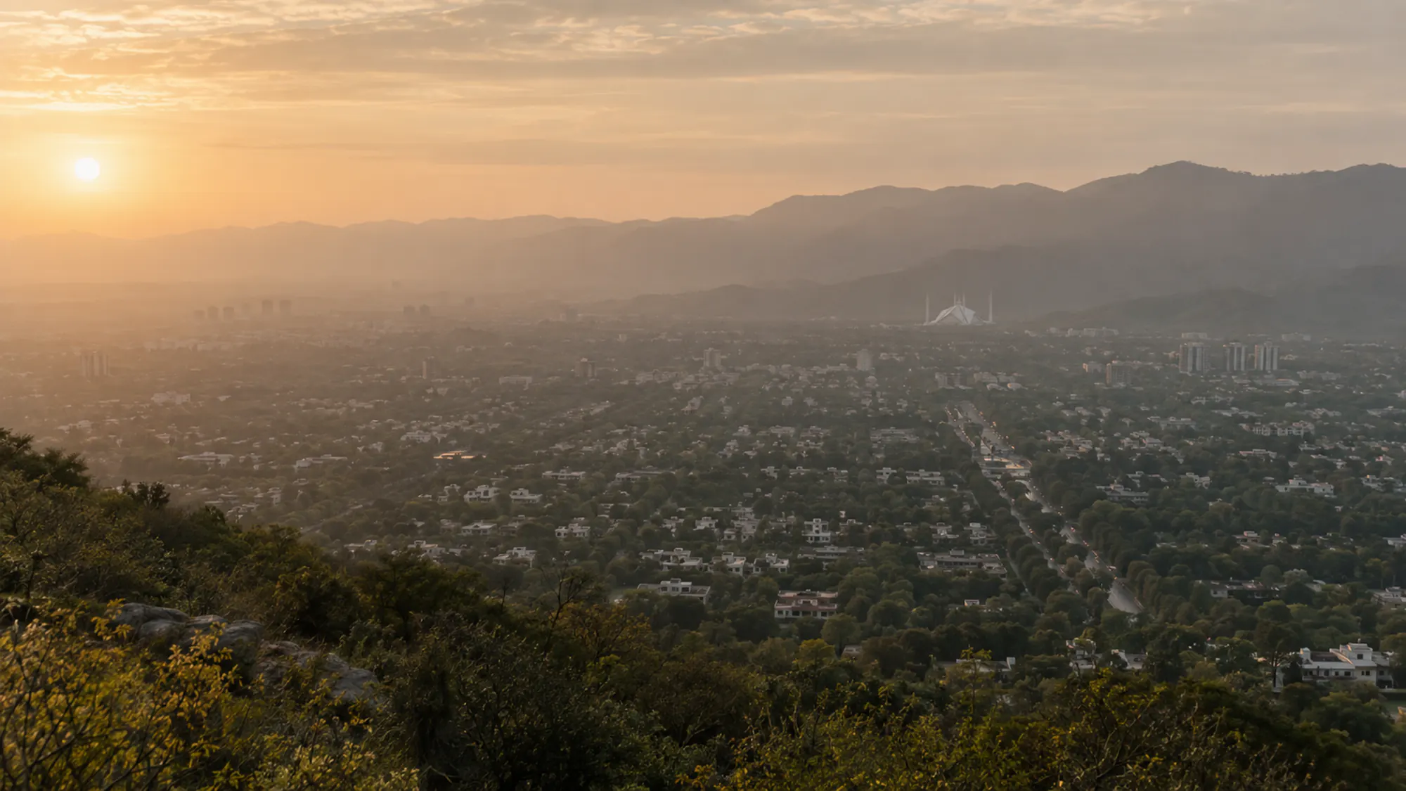 Islamabad at sunrise seen from the Margalla Hills: tree-lined sectors and low white houses stretching to the Faisal Mosque, with haze over the ridge behind.