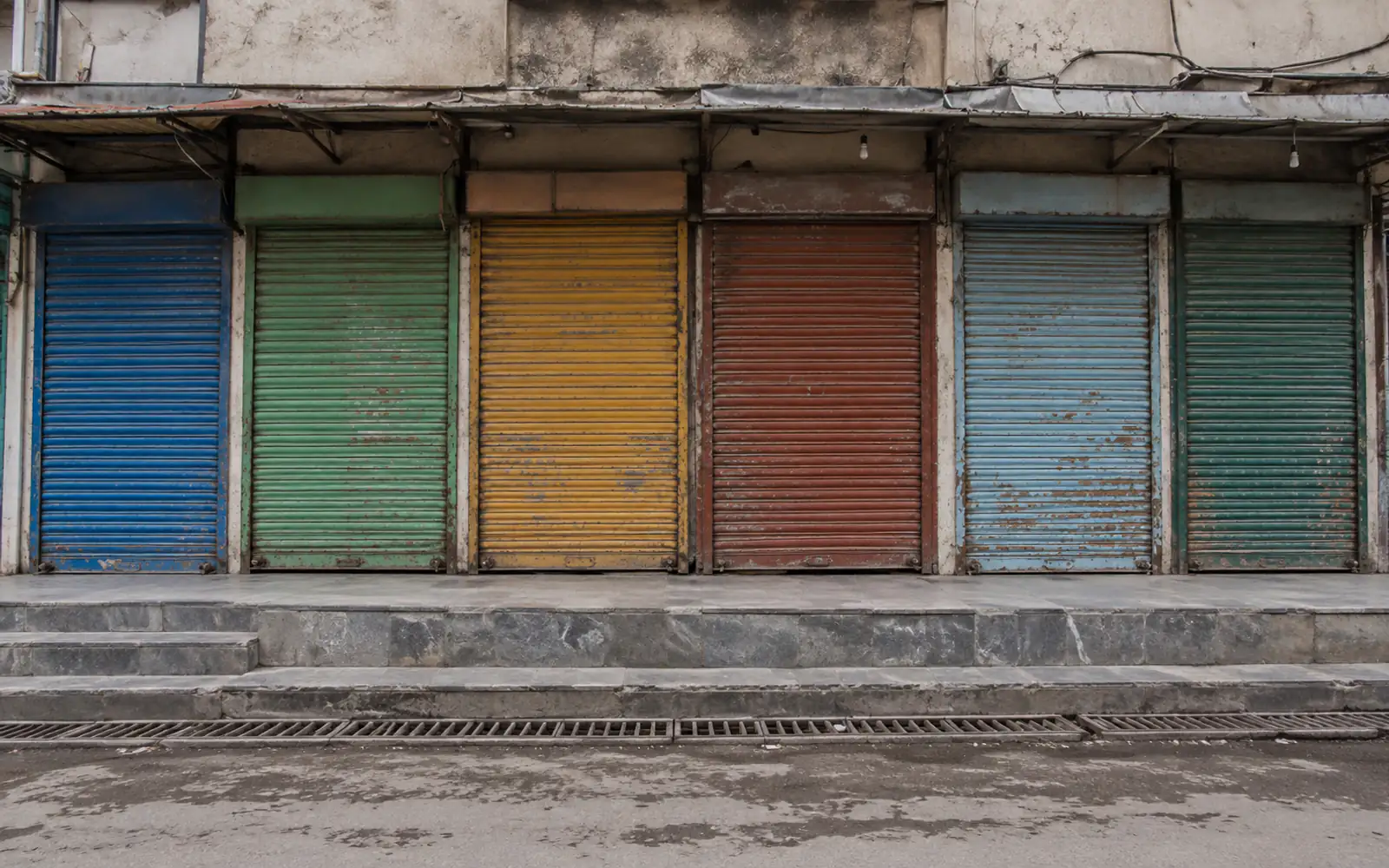 A row of shuttered shopfronts in Rawalpindi's Commercial Market, each roller shutter painted a different colour — blue, green, yellow, red — above a marble step.