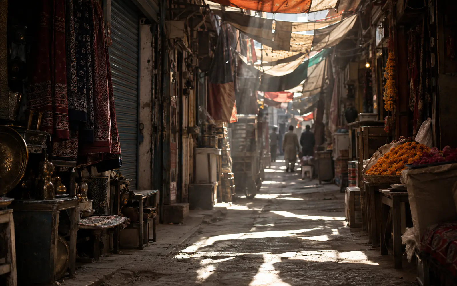 A narrow Raja Bazaar lane in Rawalpindi under strung awnings, with hanging block-printed cloth, brassware on stools, baskets of marigolds and two men walking away into the light.