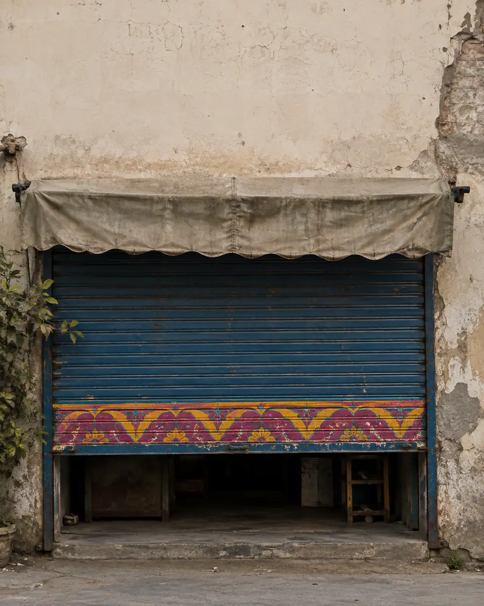 A half-raised blue shop shutter painted with a pink and yellow truck-art border, under a canvas awning on a weathered plaster wall.