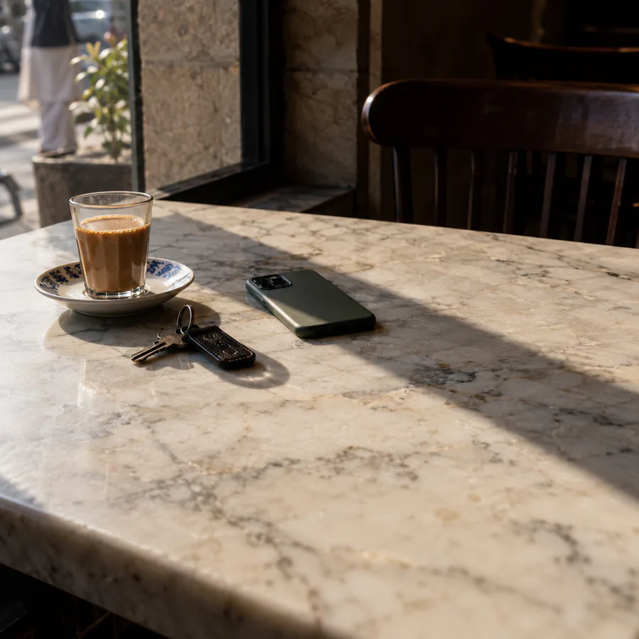 A sunlit marble cafe table with a glass of doodh patti on a blue-patterned saucer, a phone lying face down and a set of keys on a leather fob, a street visible through the window behind.